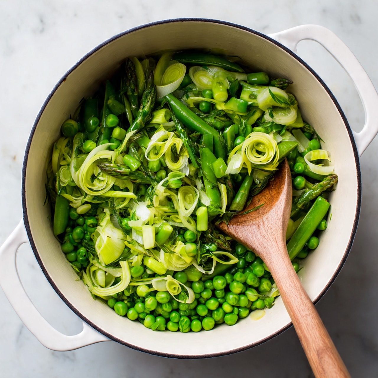 A white pot filled with a mix of bright green vegetables including snap peas, chopped asparagus, green peas, and thinly sliced leeks layered on top, showing a fresh and vibrant texture. A wooden spoon rests inside the pot on the right side. The pot sits on a white marbled surface. photo taken with an iphone --ar 4:5 --v 7