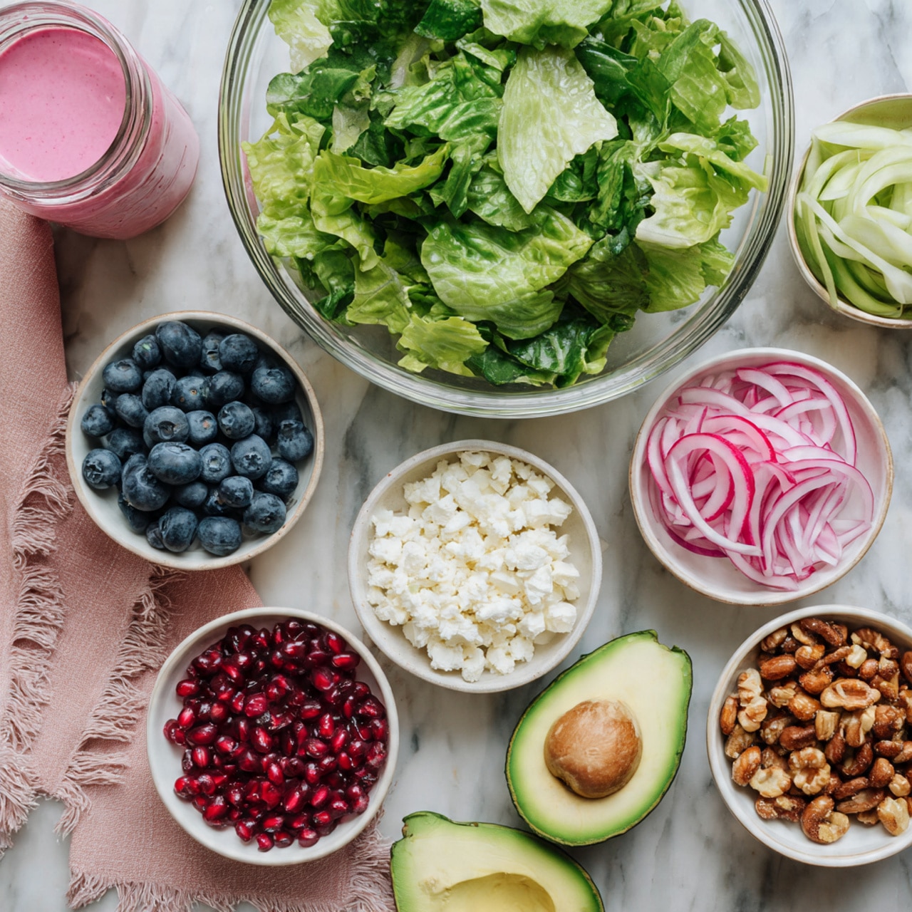 The image shows a large clear glass bowl filled with a mix of fresh green leafy lettuce placed at the top center. Below this, there are seven small white bowls arranged around the bowl of lettuce, each containing different ingredients: one holds bright blue blueberries, another has thin slices of pink and green radishes, one is filled with deep red pomegranate seeds, another contains thinly sliced light purple onions, a small bowl is filled with white crumbled cheese, and another white bowl contains roasted nuts that are golden brown and slightly shiny. To the right side are two halves of a bright green avocado with a brown seed in one half, placed on a light pink cloth with frayed edges. At the top left of the image, a small glass jar contains a thick pink sauce. All of this is on a white marbled surface photo taken with an iphone --ar 4:5 --v 7