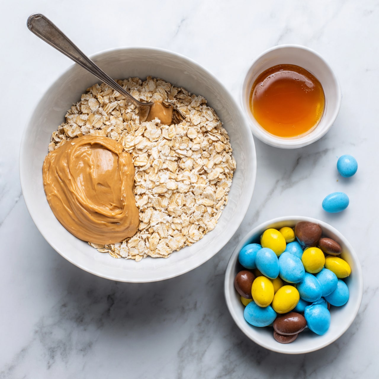 A white bowl on a white marbled surface holds three clear layers: the bottom layer is a bed of light beige oats evenly spread, on one side covered with a smooth, thick layer of light brown peanut butter, and on the other side a glossy dark amber honey layer. A spoon partially dips into the honey side. Next to the bowl, on the right, is a smaller white bowl filled with broken pieces of candy-coated chocolate eggs in pastel colors like yellow, blue, and brown. Photo taken with an iphone --ar 4:5 --v 7