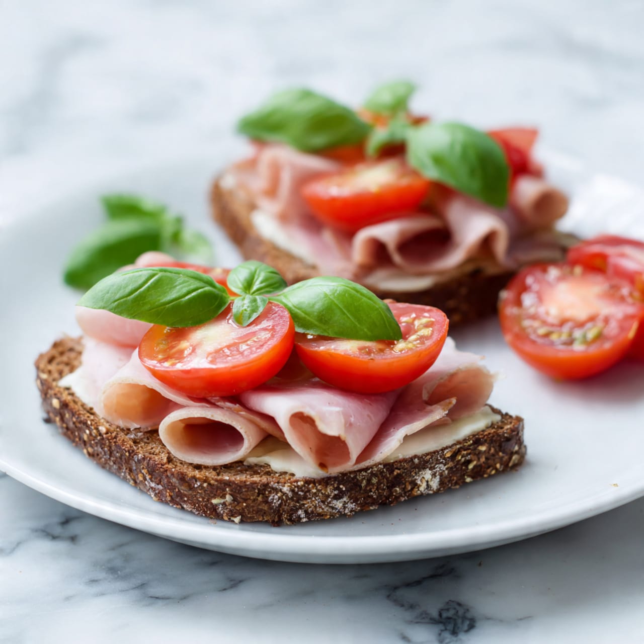 Two slices of dark rye bread are on a white plate with layers of thin pale pink deli ham folded on top of each slice. Each slice of bread also has a bright red tomato slice topped with a fresh green basil leaf. Extra slices of ham and tomato are partially visible on the side of the plate. The plate is placed on a white marbled surface. photo taken with an iphone --ar 4:5 --v 7