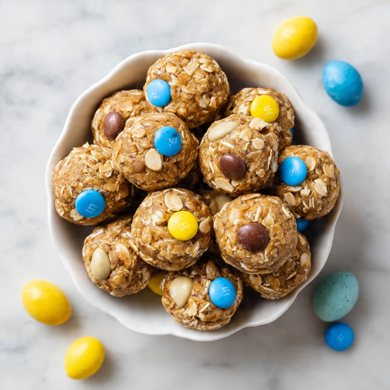 A white wavy-edged bowl filled with about two dozen round energy balls, each ball light brown with visible oat flakes and pieces of chocolate and small candy-coated chocolate eggs in yellow, blue, and pastel colors mixed in and on top, creating a textured and colorful look. The balls are slightly shiny and packed closely together, with a few loose candy-coated eggs scattered around the bowl edges. The bowl sits on a white marbled surface. photo taken with an iphone --ar 4:5 --v 7