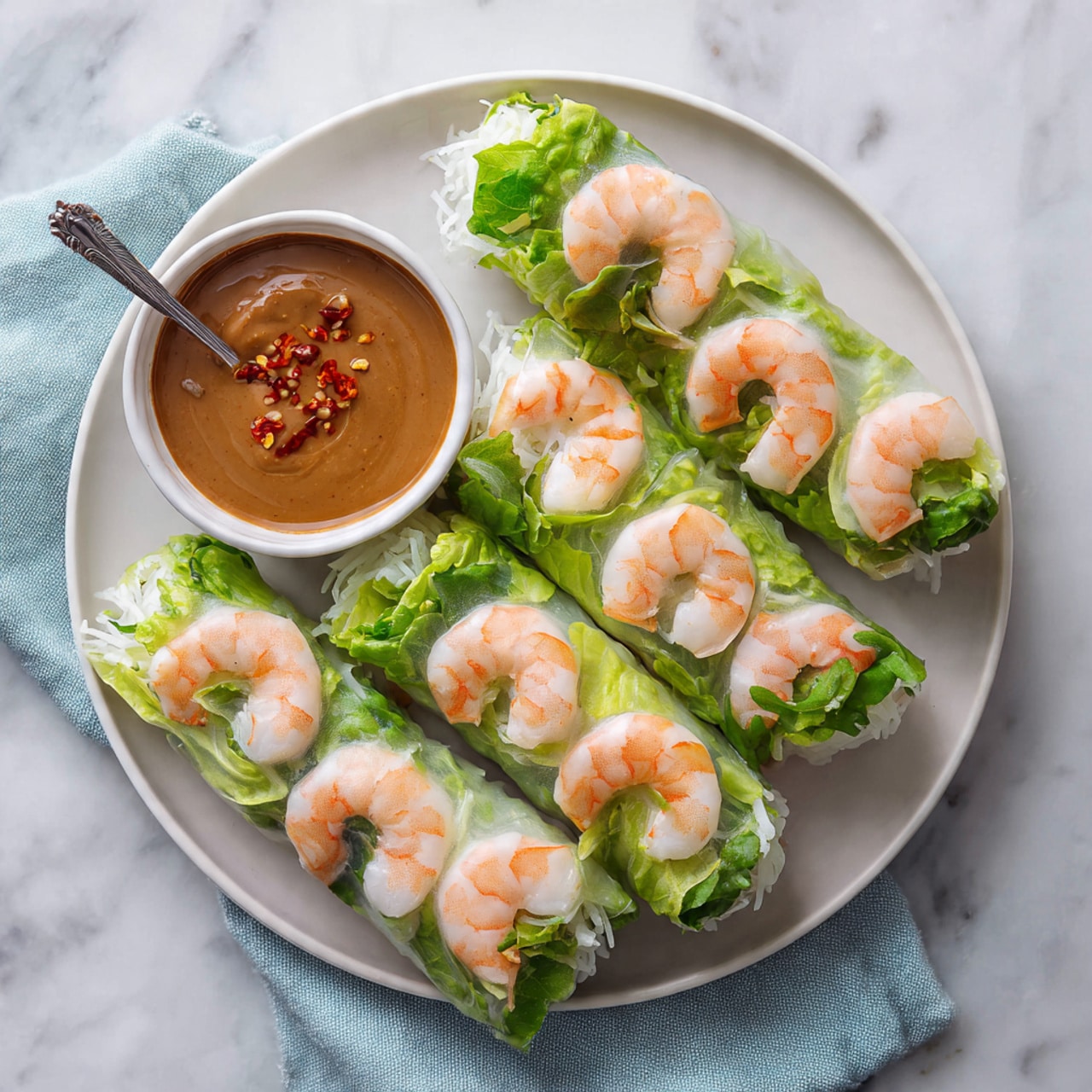 There are six fresh spring rolls on a white round plate, each roll showing a layer of translucent rice paper wrapped tightly around bright green lettuce and fresh herbs. On the outside, slightly pink cooked shrimp pieces are evenly placed in a single row along the length of each roll, their smooth texture and subtle orange stripes visible through the rice paper. Next to the rolls is a small white bowl filled with creamy brown peanut sauce, topped with a drizzle of red chili oil and a small silver spoon inside. The plate sits on a light blue cloth on a white marbled surface. photo taken with an iphone --ar 4:5 --v 7