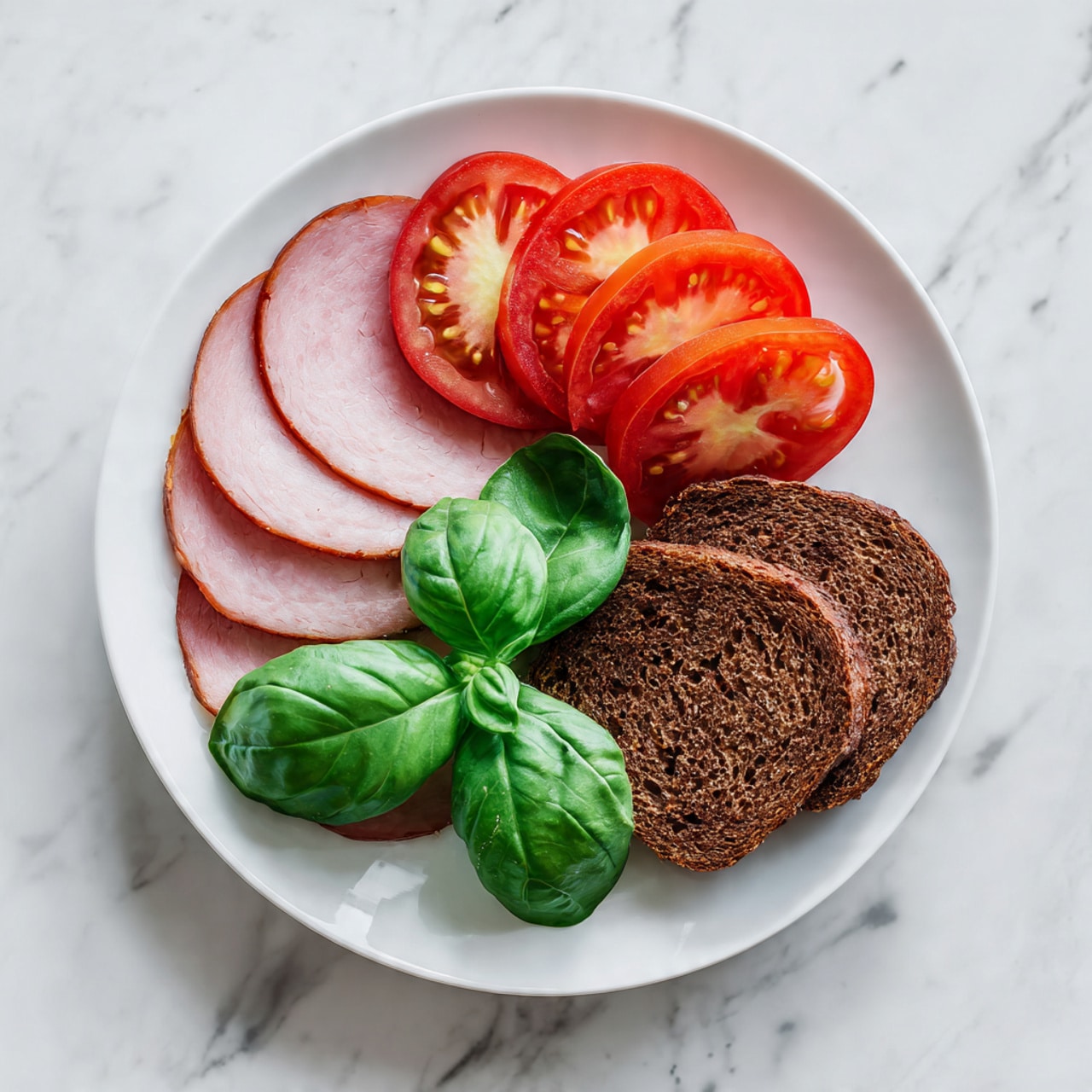 A white plate sits on a white marbled surface, holding layers of food neatly arranged. On one side, there are six fresh green basil leaves forming a curved base. Above the basil leaves, there are six thin slices of bright red tomato arranged in a slightly overlapping semi-circle. On the plate's left side, two wide, smooth slices of light pink deli ham are laid flat, partially covering the basil leaves. On the right, two round slices of dark brown, dense textured bread are placed next to the ham and tomatoes. The whole dish looks fresh and simple. photo taken with an iphone --ar 4:5 --v 7