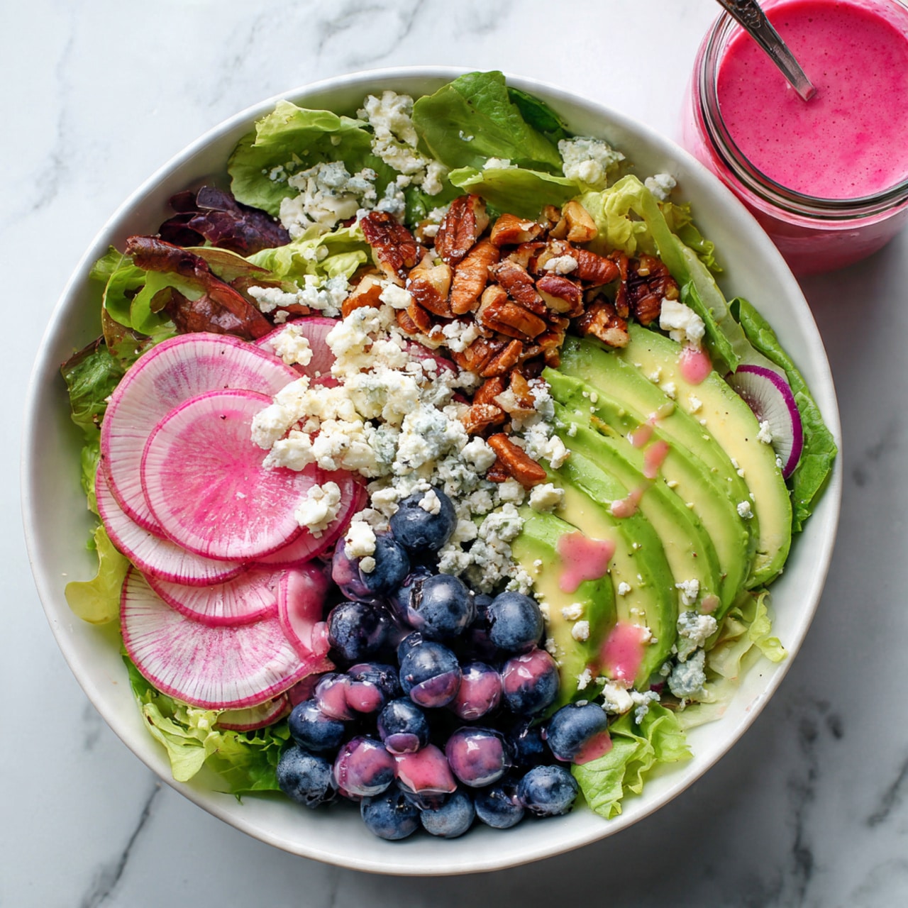 A fresh salad is served in a white bowl on a white marbled surface. The base layer is a mix of green leafy lettuce. On top, there are thin slices of watermelon radish with pink centers and light green edges, along with fan-shaped slices of bright green avocado. Scattered among these are deep blue blueberries, bright red pomegranate seeds, and toasted brown nuts. Light purple onion rings are spread evenly, and crumbled white cheese is sprinkled over the salad. A pink dressing is drizzled across the entire dish, adding a vibrant touch. A clear glass jar with the same pink dressing is placed nearby. Photo taken with an iphone --ar 4:5 --v 7