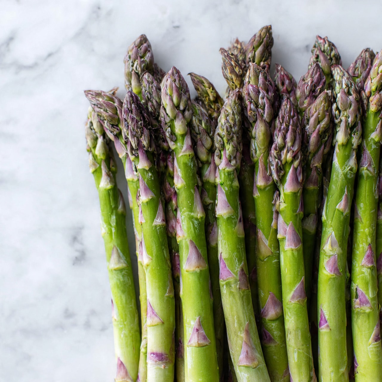 A bunch of fresh asparagus spears are arranged side by side, filling the left half of the image in a neat layer. The asparagus stalks are green with purple-tinged tips that show a tight, slightly textured surface. The spears display small triangular leaf scales along their length. The background is a smooth white marbled texture that fades into the empty space on the right side of the image. The photo is taken with an iphone --ar 4:5 --v 7