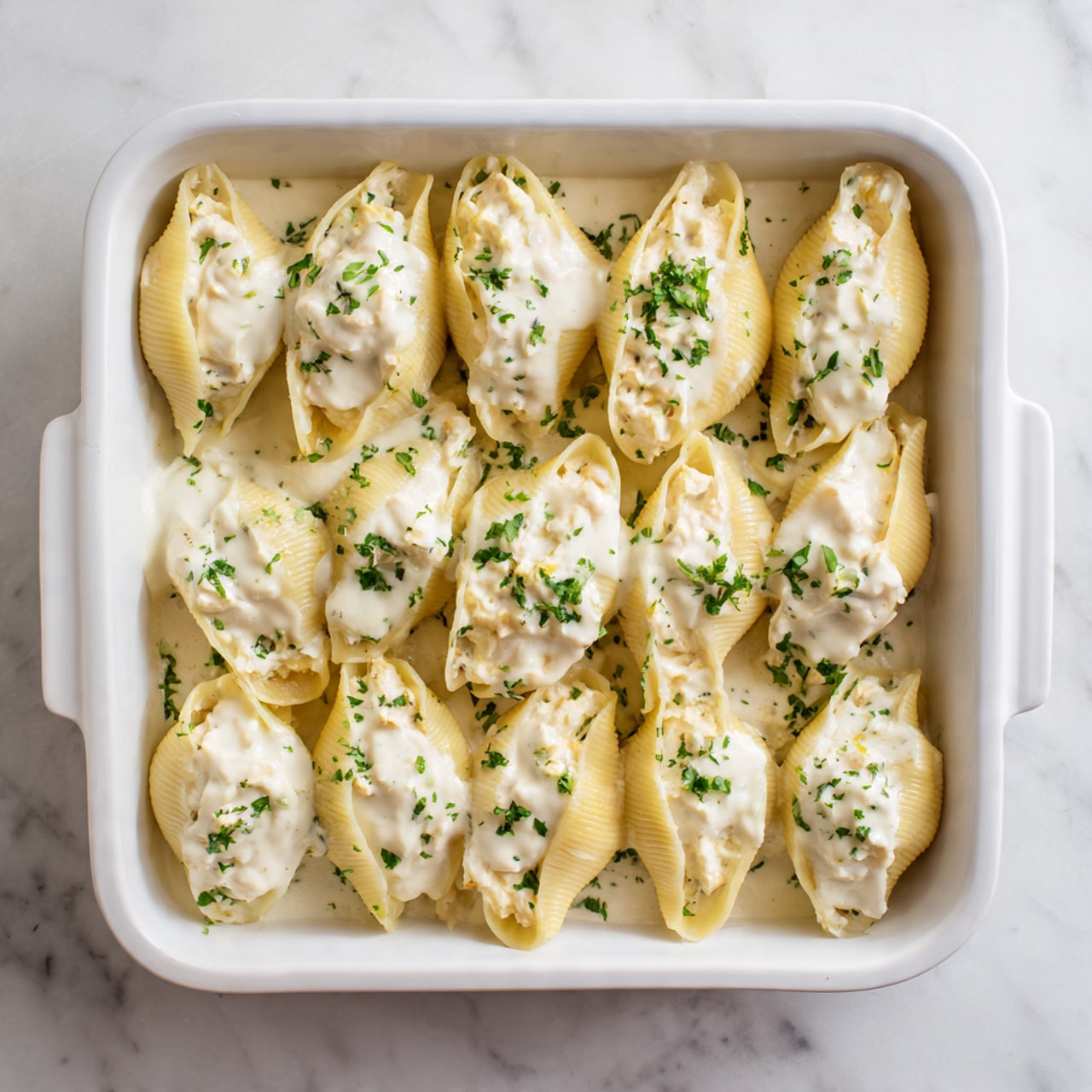 The image shows a white rectangular baking dish filled with large pasta shells. Each shell is stuffed with a light-colored filling and topped with a smooth, creamy white sauce. The shells are arranged closely together, covering the bottom of the dish in a single layer. The baking dish is placed on a white marbled surface. The overall look is creamy and soft with a pale yellow and white color palette. Photo taken with an iphone --ar 4:5 --v 7