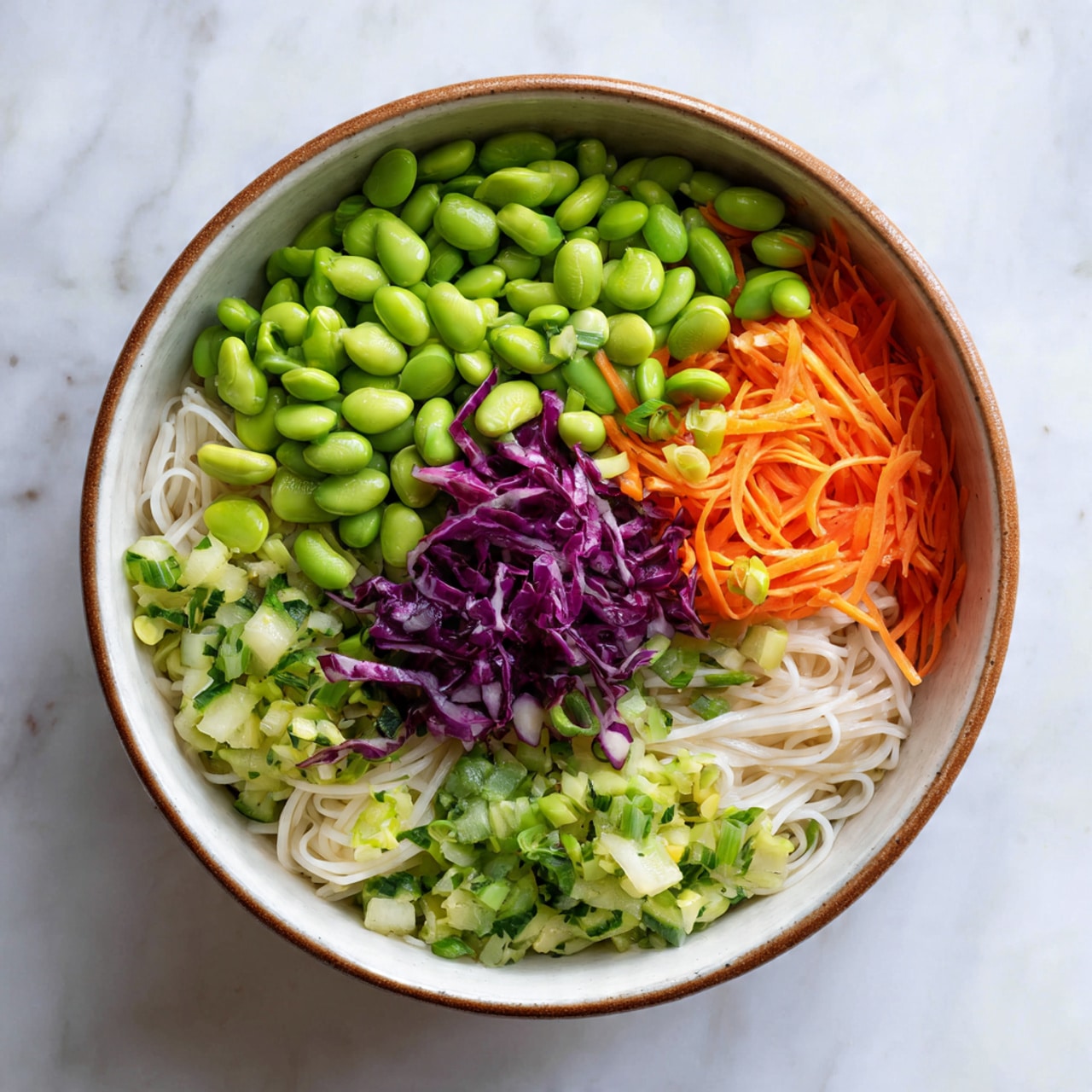 A bowl filled with a colorful noodle salad showing three main layers: the bottom layer has thin white noodles spread throughout, the middle layer is mixed with light green chopped lettuce and cucumber pieces, and the top layer includes bright green edamame beans, thin orange carrot sticks, and small bits of purple cabbage, all mixed evenly. The bowl is white with a brown rim, resting on a white marbled surface. Photo taken with an iphone --ar 4:5 --v 7