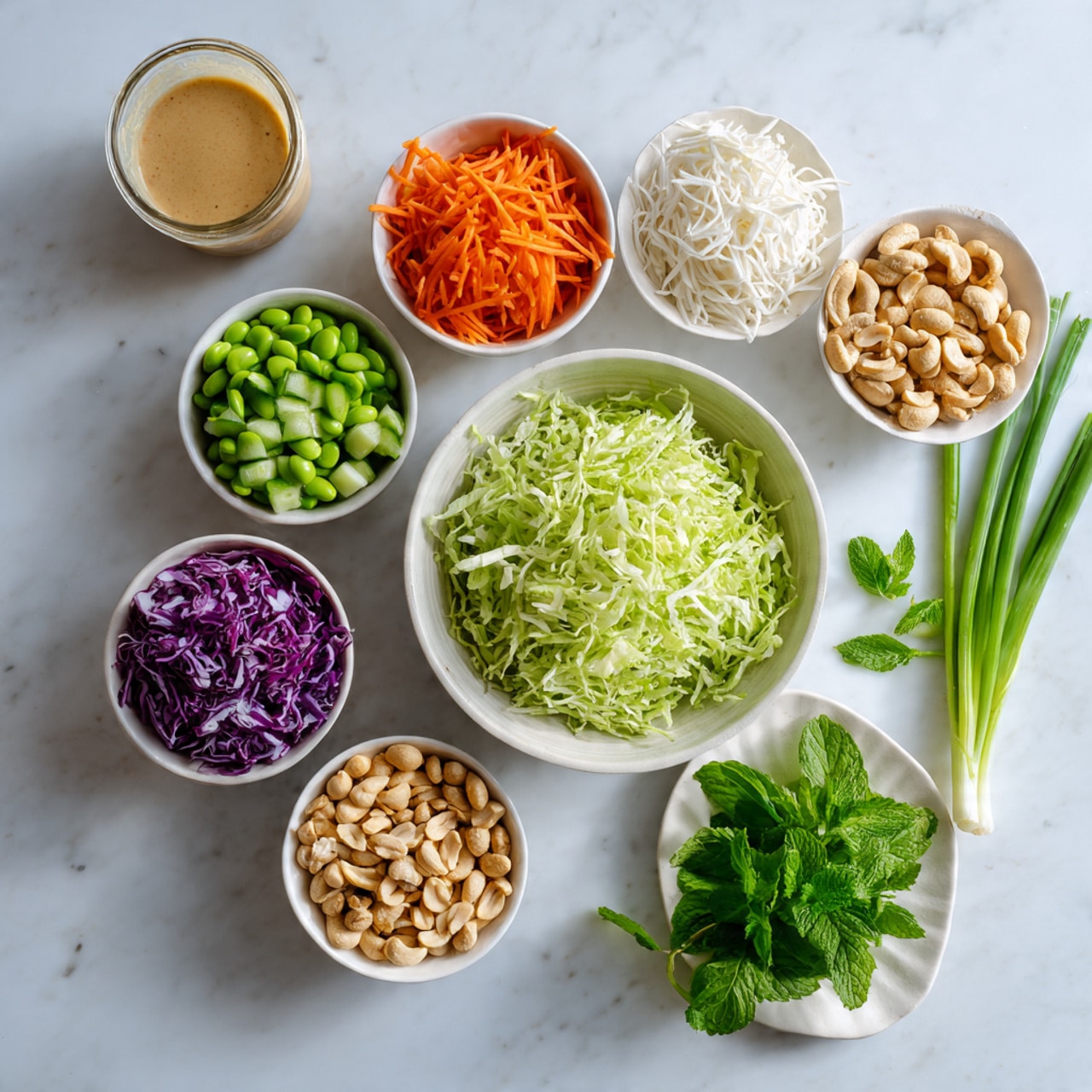 This image shows a clean white marbled surface with several white bowls and one small white plate arranged neatly. The largest bowl is filled with shredded light green lettuce, placed near the center. Surrounding it are smaller white bowls containing bright orange shredded carrots, thin white noodle strands, chopped green cucumber pieces, vibrant green edamame beans, purple shredded cabbage, and a mix of light brown peanuts and cashews. To the right side, a small white plate holds fresh green herbs including mint and cilantro. Two long green onions lie beside the plate. A small glass bowl with a creamy light brown dressing is at the top left corner. The setup is bright and fresh, centered in the frame with soft lighting. Photo taken with an iphone --ar 4:5 --v 7