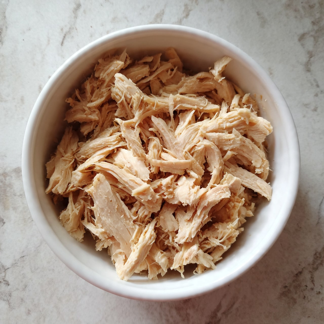 A white bowl filled with shredded cooked chicken, showing many pieces in light beige color with soft, fibrous texture. The chicken pieces are arranged loosely inside the bowl on a white marbled surface. Photo taken with an iphone --ar 4:5 --v 7