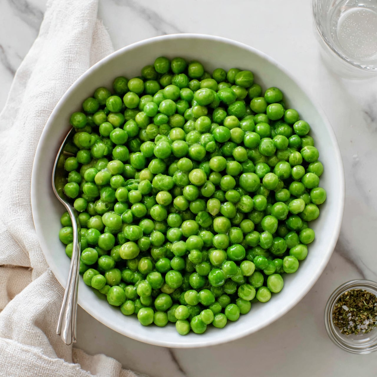 A white bowl filled with bright green peas that look fresh and slightly shiny, showing their round and smooth texture. The peas fill the bowl almost to the top in one thick layer. A silver spoon with a simple design is placed inside the bowl on the left side, partly covered by the peas. The bowl sits on a white marbled surface with a soft white cloth nearby and a small clear container of seasoning visible on the lower right side. Photo taken with an iphone --ar 4:5 --v 7