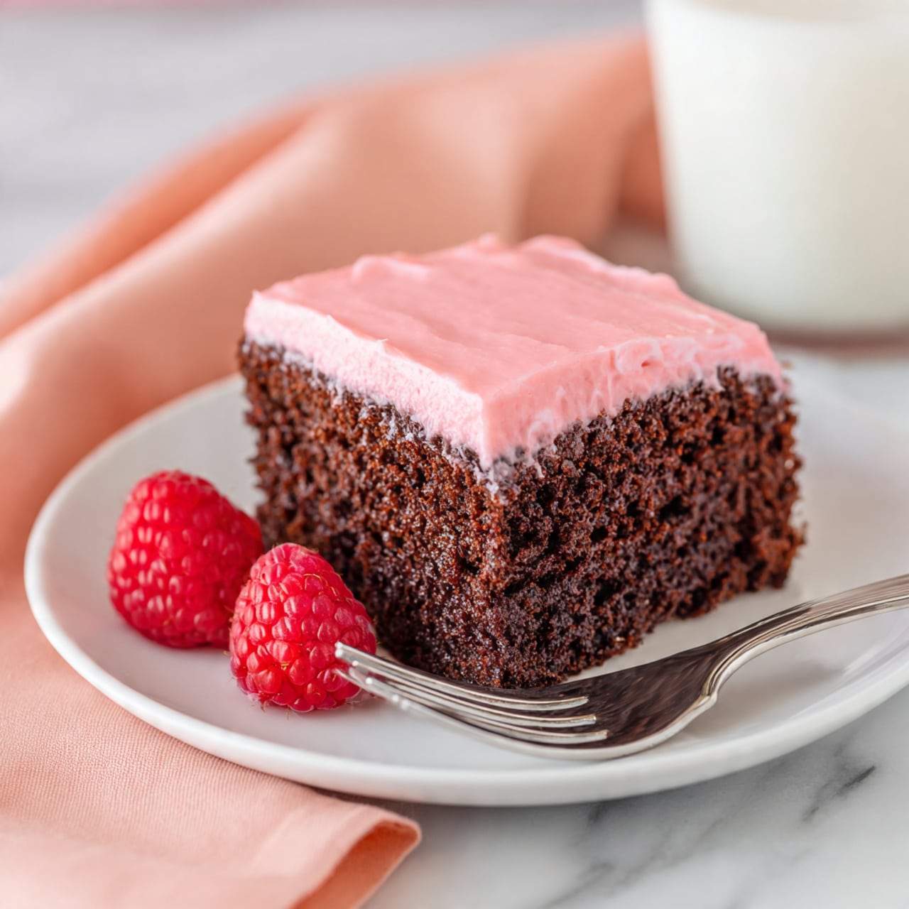A piece of chocolate cake with one thick layer is shown on a white plate. The cake layer is dark brown with a moist texture, topped with a smooth, evenly spread bright pink frosting. On the plate near the cake are two fresh red raspberries. A silver fork holds a small bite of the cake with frosting next to the cake piece. The setting is on a white marbled surface with a soft peach napkin and a glass of milk blurred in the background. Photo taken with an iphone --ar 4:5 --v 7