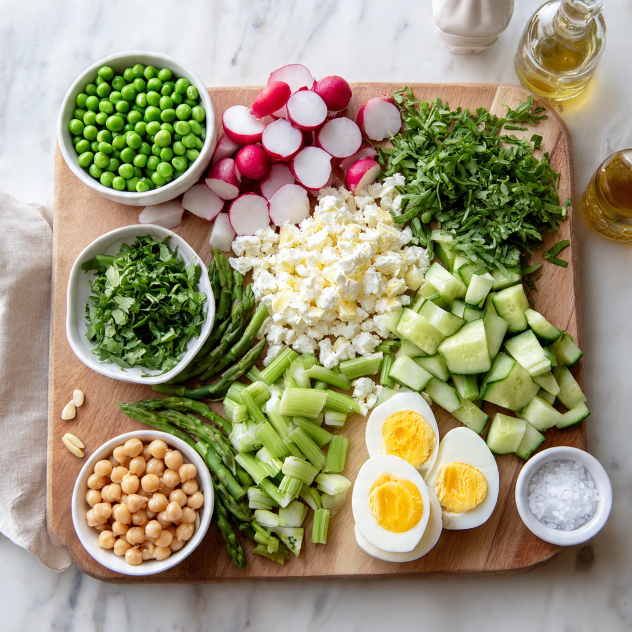 The image shows a wooden board filled with fresh ingredients arranged in neat piles. There are bright green peas in a small white bowl on the left, next to chopped radishes with white centers and red edges. In the middle, there is a heap of crumbly white cheese, some green asparagus pieces, and chopped green herbs. To the right, there are halved boiled eggs with bright yellow yolks, sliced cucumber with light green skin, chopped herbs, and a white bowl with golden chickpeas. Another small white bowl holds light brown pine nuts. The board sits on a white marbled surface with a salt container and a bottle of dressing nearby. photo taken with an iphone --ar 4:5 --v 7
