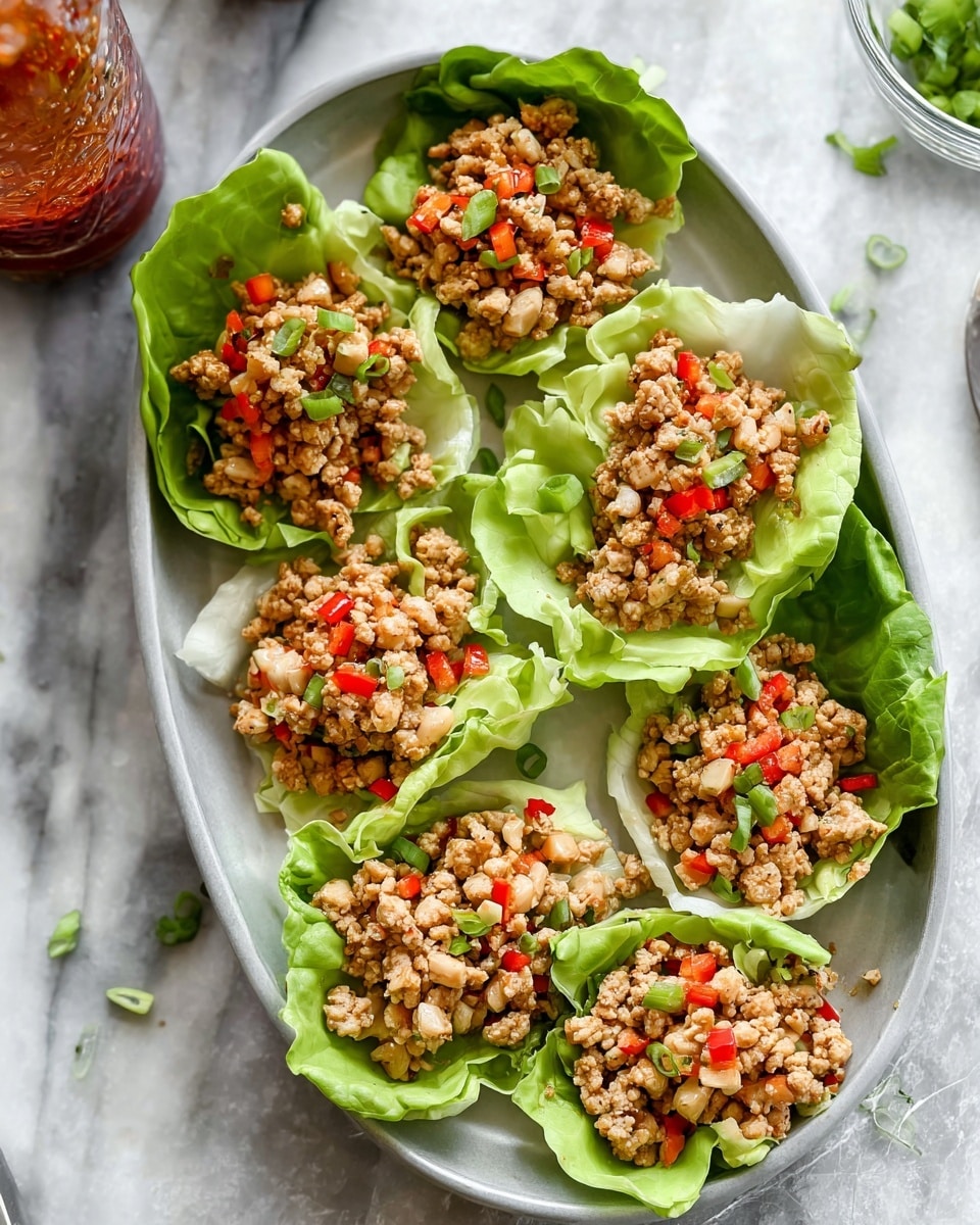 The image shows six lettuce cups on a white oval plate, each cup made with green leafy lettuce wrapped around finely chopped cooked meat mixed with small red and green bell peppers and diced pale vegetables. The meat mixture is light brown and crumbly, filling each lettuce cup fully and forming the top layer. The plate is set on a white marbled surface with a glass bottle with some red sauce seen at the upper left corner and chopped green onions scattered near the plate. Photo taken with an iphone --ar 4:5 --v 7